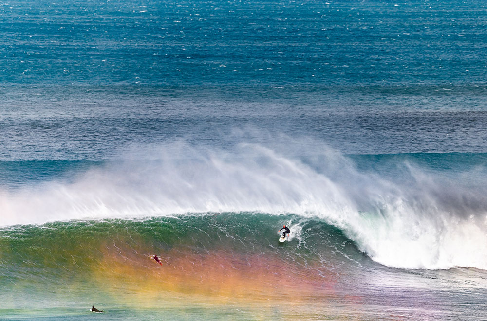 A surfer rides a large wave as a rainbow forms in front of him. 